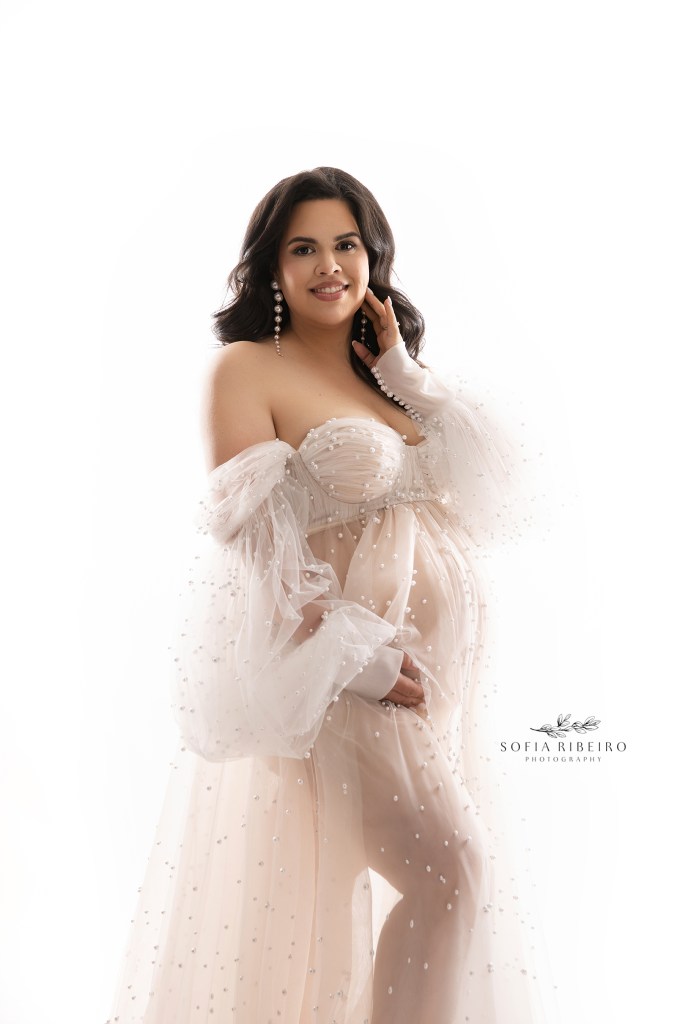 mom poses to the camera, holding her baby bump in a sheer pearl gown