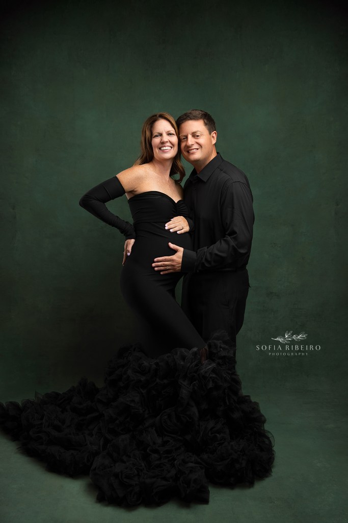 mom and dad to be posing in matching black outfits against a green backdrop for maternity photos in nj